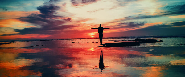 A person standing on a rock in the water with arms outstretched facing the sunrise in the pink clouded sky
