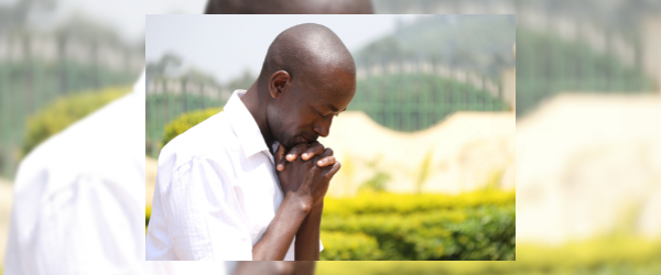 Close up of a Kenyan man with his eyes closed, head bowed, and hands clasped in a prayer position outside in front of green plants