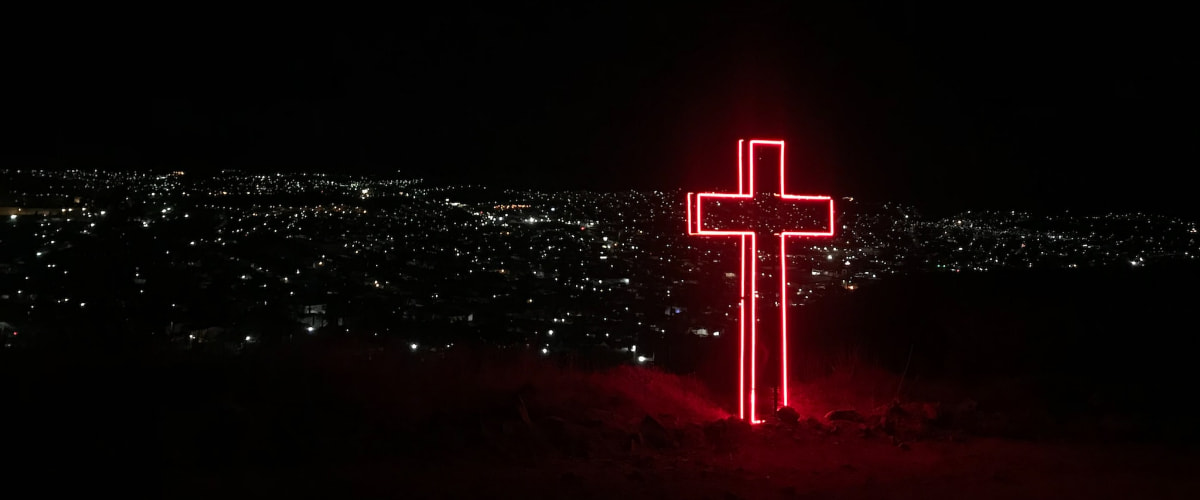 Glowing Neon Red Cross sitting atop a hill overlooking town lights at night
